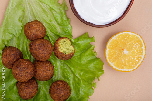 Close up with fried falafel on salad leaf, white yogurt sauce in clay bowl and fresh yellow lemon on beige background. Healthy roasted vegetarian fastfood. Israeli cuisine.