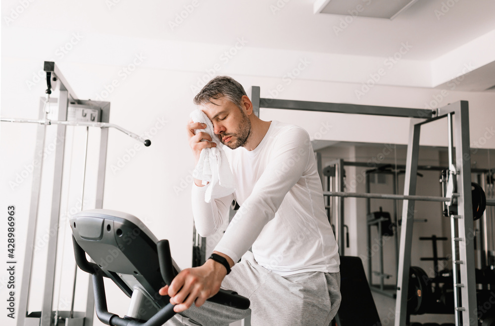a tired man wipes sweat from his face with a towel during an exercise on an exercise bike. a tired man wipes sweat from his face with a towel during an exercise on an exercise bike.