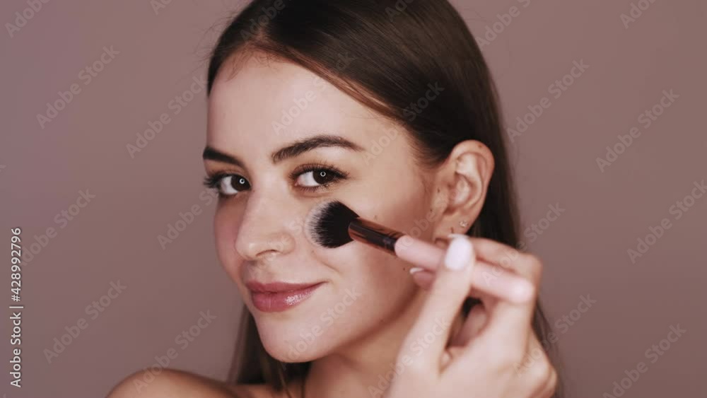 Natural makeup. Beauty skincare. Cosmetic product. Portrait of satisfied brunette woman enjoying applying powder using face brush isolated on brown background. Natural makeup. Beauty skincare. Cosmetic product. Portrait of satisfied brunette woman enjoying applying powder using face brush isolated on brown background.