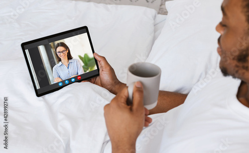 Young African Man Relaxing In Bed With Digital Tablet, Making Video Call
