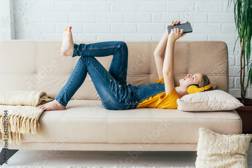 Teenage girl in denim overalls relaxes in pleasure on the couch with an electronic tablet. The child communicates in video chat with wireless Internet devices.