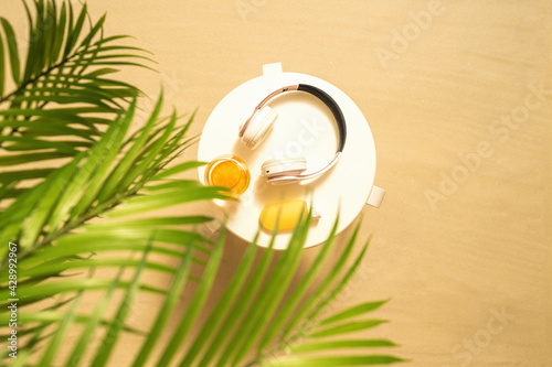 Headphones, sunscreen, orange juice on the white table on the sandy beach in the palm tree forest. Summer vacation. Top view. Concept