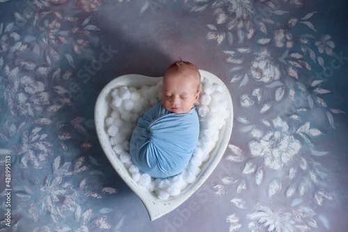 Newborn baby boy wrapped in wrap on sleep in wooden bowl in the shape of heart with natural props.
