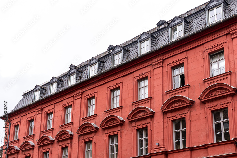 Obraz premium Vibrant red stone tenement or apartment building with shiny black roof tiles, attic windows (dormers), many windows with geometric ornament and triangle pediments.