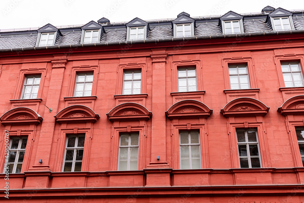 Obraz premium Vibrant red stone tenement or apartment building with shiny black roof tiles, attic windows (dormers), many windows with geometric ornament and triangle pediments.