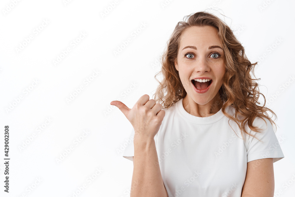 Excited blond girl with amazed face, gasping and looking at camera, pointing finger aside, showing promo on copy space, standing over white background Excited blond girl with amazed face, gasping and looking at camera, pointing finger aside, showing promo on copy space, standing over white background