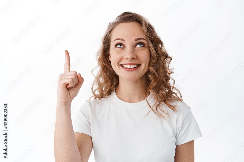 Young blond woman looking and pointing up with pleased smile, reading promotional text, showing advertisement, white background Young blond woman looking and pointing up with pleased smile, reading promotional text, showing advertisement, white background