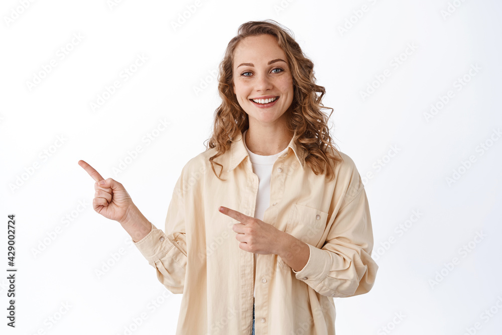 Young attractive woman smiling and demonstrating promo text, pointing fingers left at copyspace, showing advertisement, standing over white background