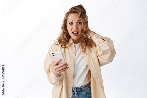 Woman holding phone and look in panic, feeling anxious and worried about something posted online, standing with smartphone against white background