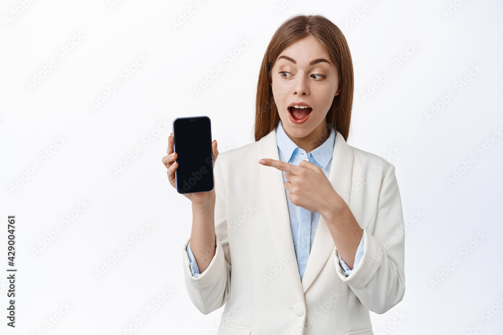 Excited girl in business suit, points at mobile screen and looks amazed, checking out new app feature, showing advertisement on display, standing over white background Excited girl in business suit, points at mobile screen and looks amazed, checking out new app feature, showing advertisement on display, standing over white background