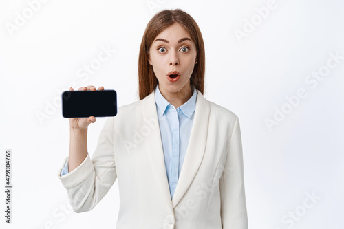 Amazed ceo office woman in business suit shows mobile screen, holding phone horizontally and demontrate empy display, white background photo