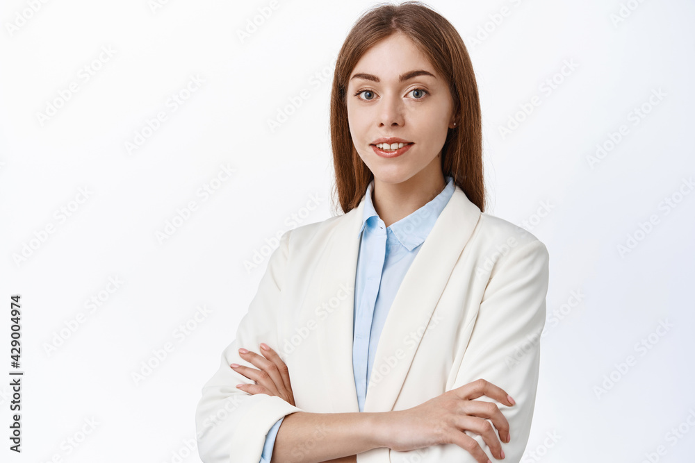 Confident young businesswoman in suit, cross arms on chest and smile at camera, look determined and skilfull, standing over white background Confident young businesswoman in suit, cross arms on chest and smile at camera, look determined and skilfull, standing over white background