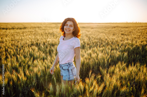 Smiling girl stands in the field. Happy woman at sunset in wheat. A girl in a pink T-shirt stands against the background of the field and the setting sun. © Konstantin Zibert