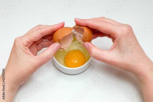 women's hands breaking a raw chicken egg into a small white cup on a white background