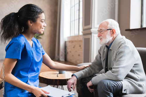 Quadro su tela Young woman physician and old mature senior man grandfather talking providing medical assistance sitting on sofa