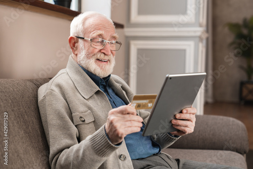 Cheerful old grandfather man doing online payment from digital tablet with credit card. Elderly senior caucasian grandpa paying bills online with credit card © InsideCreativeHouse