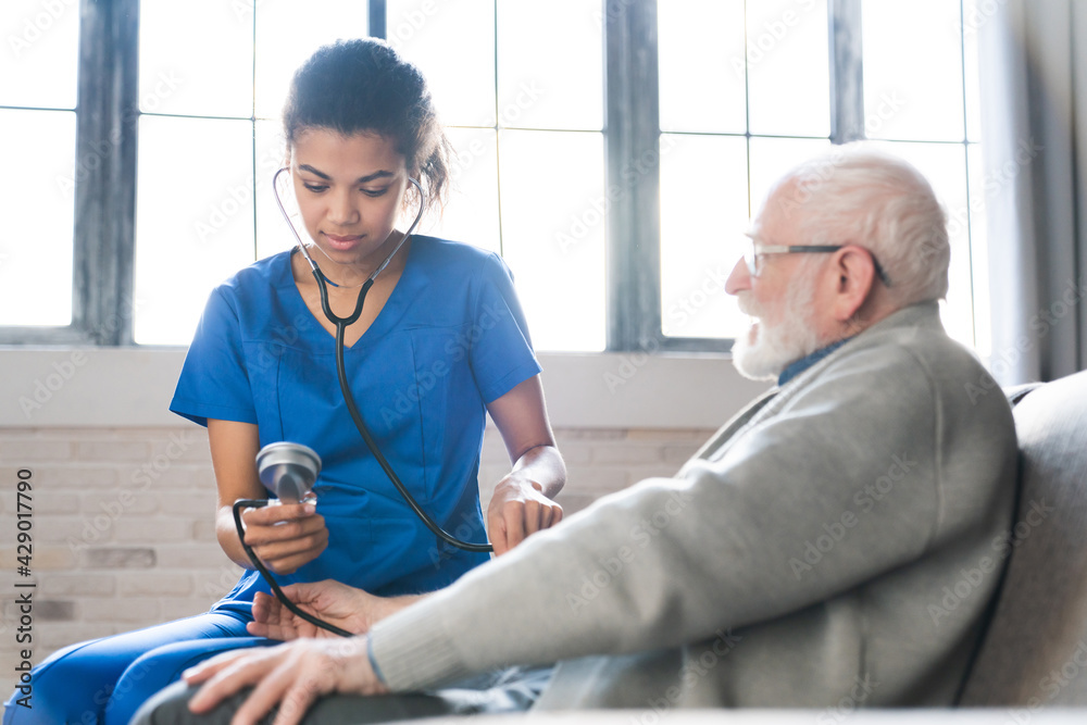 Fototapeta premium Young african nurse measuring blood pressure of elderly man indoors. Assisting senior people. Caregiver in nursing home checking blood pressure of an old retired grandfather man. Revealing shot.