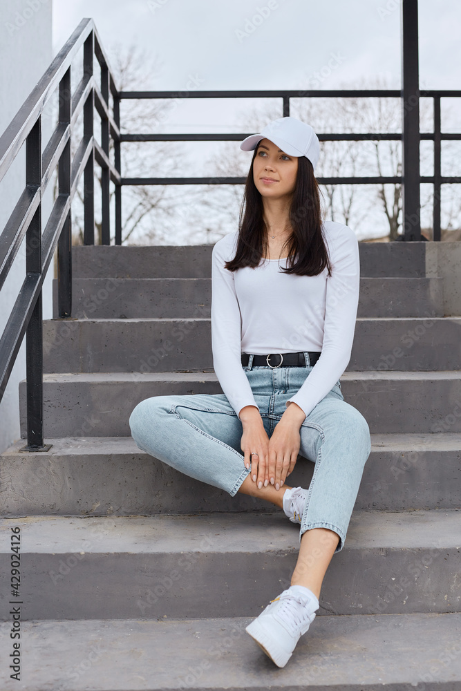 Young attractive dark haired female sitting on stairs and looking away with pensive dreamy facial expression, stylish woman wearing jeans, white shirt and baseball cap posing outdoors.