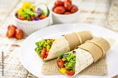 Quadro su tela Two vegetarian tortilla wraps on wooden cutting board with vegetables in the bac