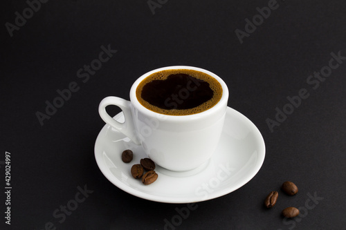 Black coffee in the white cup and coffee beans on the black background. Close-up. Copy space.