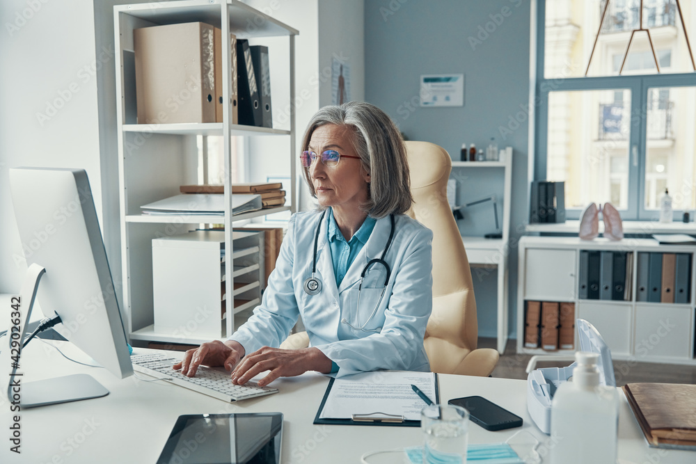 Confident mature female doctor in white lab coat working on computer while sitting in her office