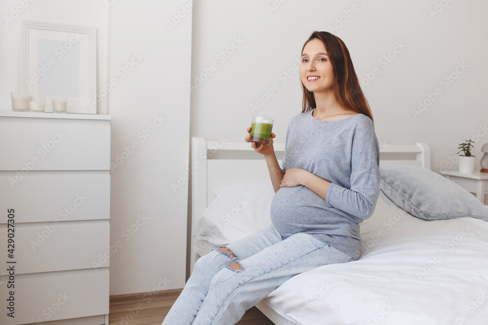 Young beautiful pregnant woman sitting on the bed with white linen and drinking a green smoothie.