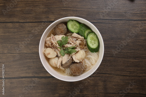 Clear white rice vermicelli beehoon noodle soup with chicken beef meat ball shredded cucumber in white bowl chopsticks spoon chili soya sauce parsley garnish