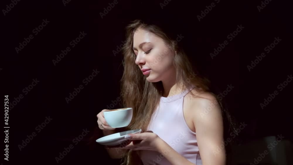 Girl with long light brown hair, grey eyes and long eyelashes dressed in short pink dress drinking coffee with pleasure from a white cup.  Medium shot from the side, filmed in a darkened room