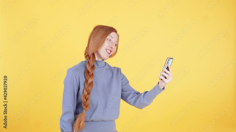 Young woman with freckles and long ginger plait is talking to someone on the phone using video call and smiling. Yellow background Young woman with freckles and long ginger plait is talking to someone on the phone using video call and smiling. Yellow background