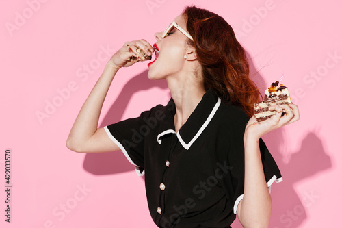 woman in a black dress and glasses eating cakes on a pink background side view