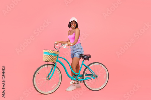 Fototapeta Naklejka Na Ścianę i Meble -  Full length of happy black lady in summer outfit standing with classic bicycle on pink studio background