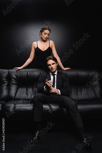 seductive woman in slip dress standing near man sitting on leather couch on black.