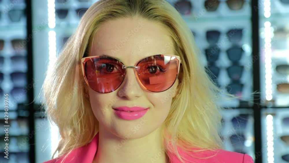 Attractive blonde woman in pink suit with elegant large sunglasses shakes hair near display in fashion optical store close view Attractive blonde woman in pink suit with elegant large sunglasses shakes hair near display in fashion optical store close view