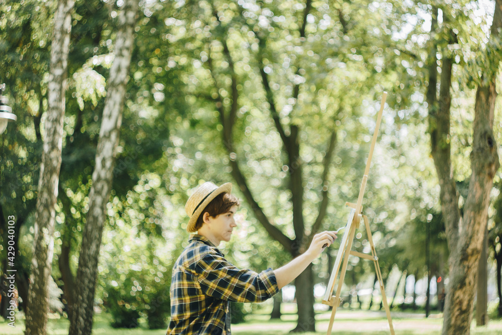a young guy in a straw hat sits in the park in front of an easel and paints a picture with oil paints. Artistic work in nature. Plein air