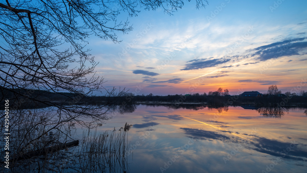 Evening landscape with a river. Village and forest on the horizon. Sunset on the river. The sky and evening clouds are reflected in the water.