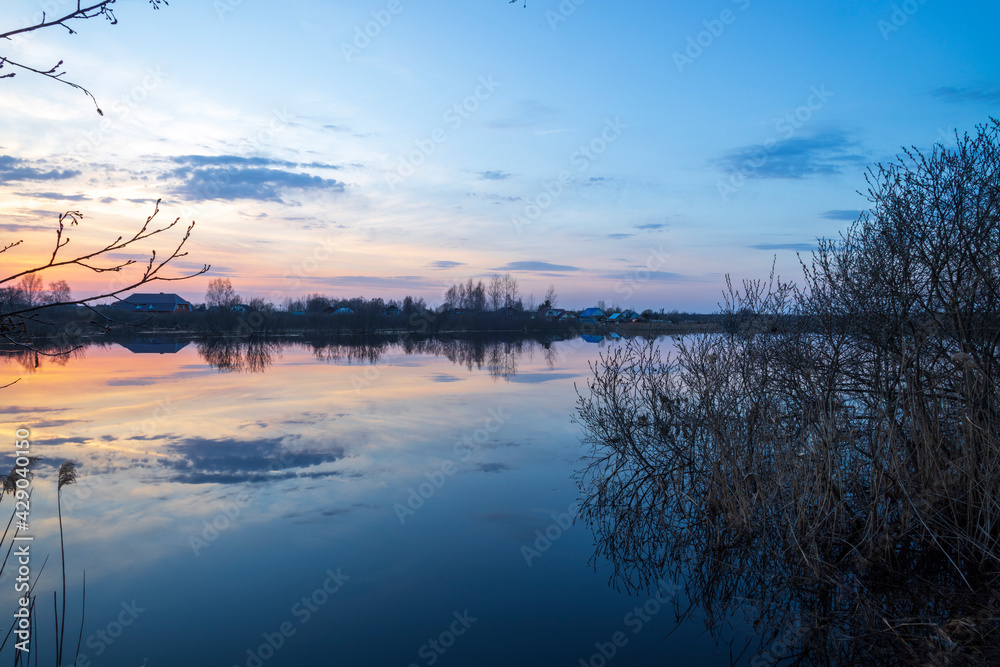 Evening landscape with a river. Village and forest on the horizon. Sunset on the river. The sky and evening clouds are reflected in the water.