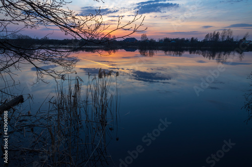 Evening landscape with a river. Village and forest on the horizon. Sunset on the river. The sky and evening clouds are reflected in the water. © Sergei