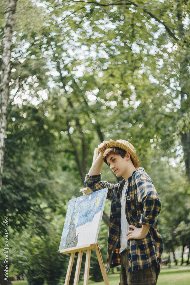 Φωτογραφία a young guy in a straw hat sits in the park in front of an easel and paints a picture with oil paints