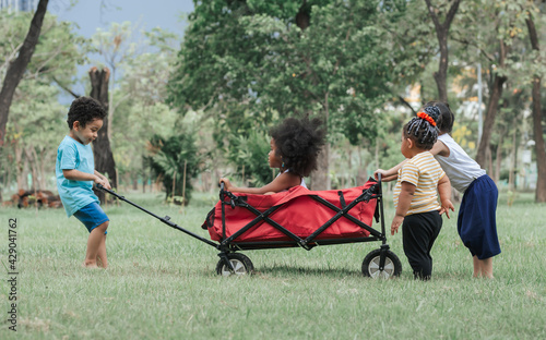 Mixed race little cute children helping each other to push and drag a red cart while playing in outdoor green park. Education, Diversity, Unity and Teamwork Concept.