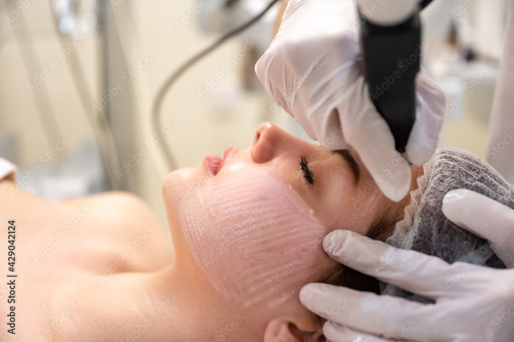 Calm young woman is getting anti-aging rejuvenation procedure at beauty salon. Beautician hands holding tool under her face.