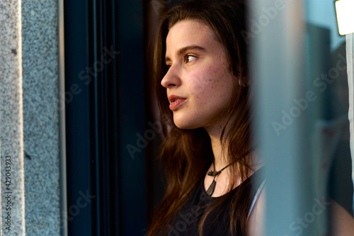 young woman with long hair and light skin, black top and pants leaning out of a window with sunset. pretty teen going out of a glass sunset. sexy caucasian girl going out to the balcony through window