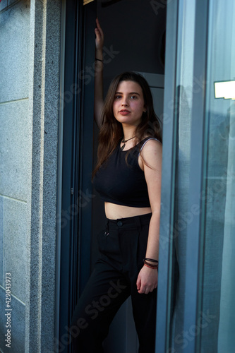 young woman with long hair and light skin, black top and pants leaning out of a window with sunset. pretty teen going out of a glass sunset. sexy caucasian girl going out to the balcony through window