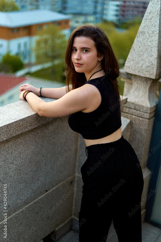 young woman with long hair and white skin in a black top and pants leaning out of the balcony of a stone building at sunset. teenager on the terrace of a happy penthouse. pretty and sexy girl