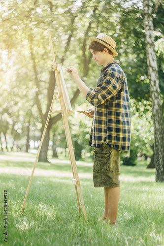 a young guy in a straw hat sits in the park in front of an easel and paints a picture with oil paints. Artistic work in nature. Plein air