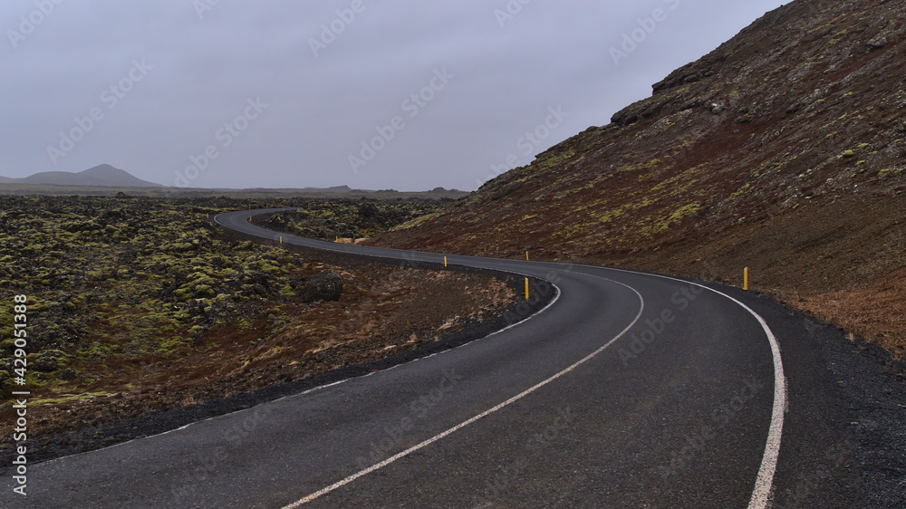 Diminishing perspective of empty winding country road (s-shape) beside moss covered lava field of volcanic stones near Grindavik, Reykjanes peninsulsa, Iceland on cloudy winter day.