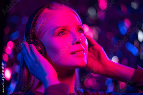 Happy young blonde woman listens to music with headphones in a nightclub. The girl smiles, dances and holds headphones with her hands. Blurred background, close-up, blue and red lights. Nightlife, fun