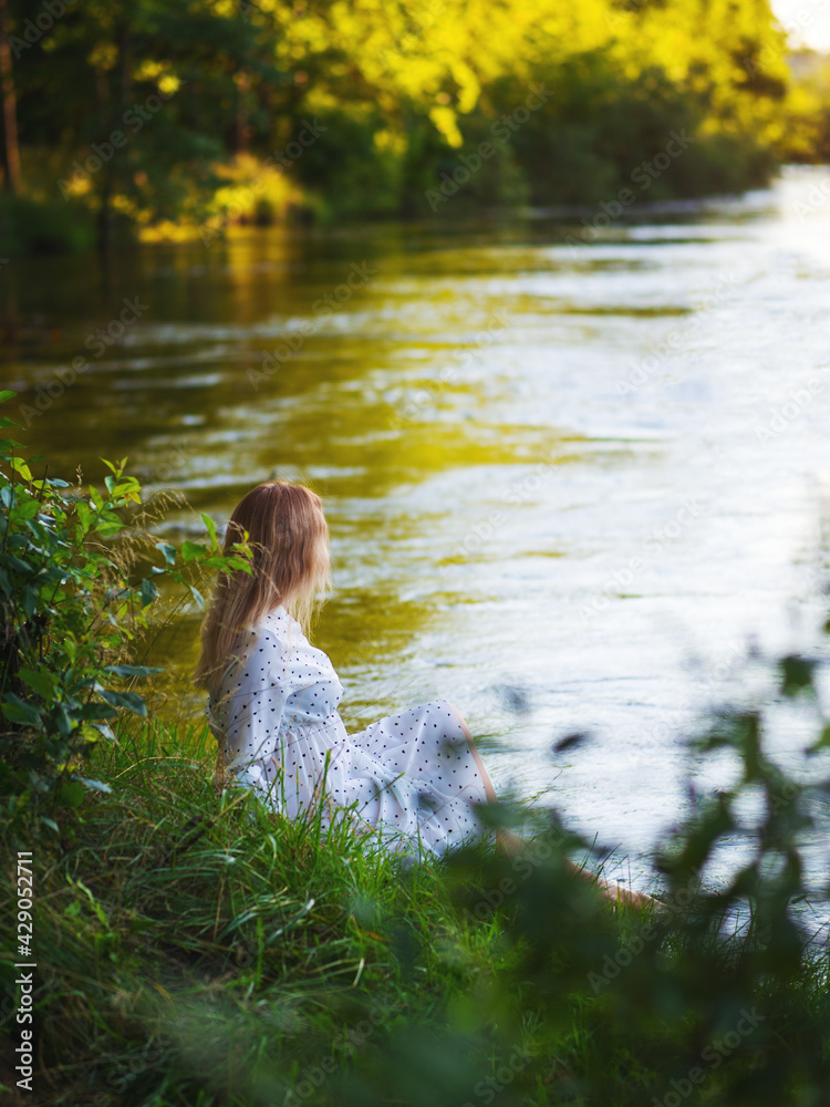 Cute beautiful young woman in white dress sitting on the river bank, sunny summer evening Cute beautiful young woman in white dress sitting on the river bank, sunny summer evening
