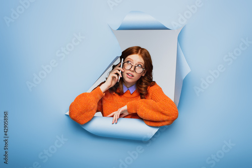 redhead young woman in orange sweater adjusting glasses and talking on cellphone on blue ripped background. © LIGHTFIELD STUDIOS