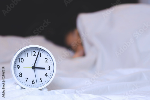 Focus at white round alarm clock in 3 o'clock with blurred background of woman is sleeping on bed at night
