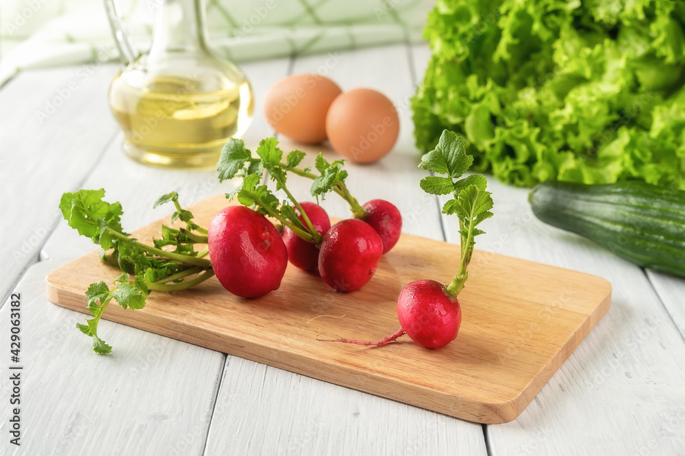 Wet whole radishes with green leaves on a wooden cutting board over a white table. Ingredients for raw spring vegetable salad with eggs. Low calories healthy eating concept. Wet whole radishes with green leaves on a wooden cutting board over a white table. Ingredients for raw spring vegetable salad with eggs. Low calories healthy eating concept.
